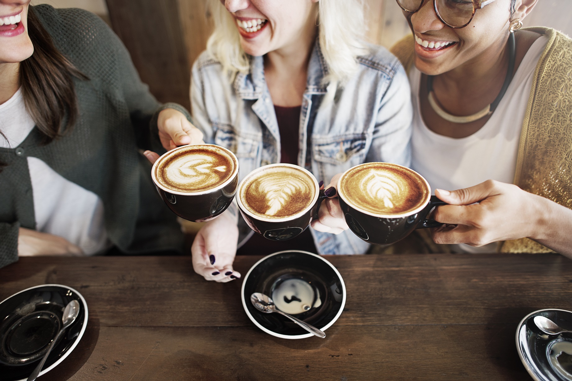 Three friends sitting together at a café, smiling and holding cups of latte art coffee over a wooden table.